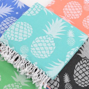 May include: A collection of colorful beach towels with a white pineapple pattern. The towels are in shades of blue, teal, coral, green, and black. Each towel has a white fringe along the edge, perfect for a day at the beach or pool.