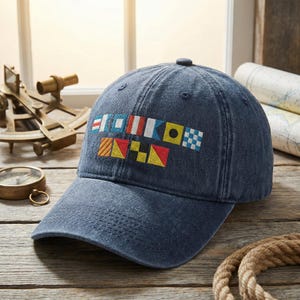 May include: A navy blue baseball cap with embroidered nautical flag designs. The cap has a curved brim and is made of a soft, worn fabric. The background includes nautical instruments and a rope.