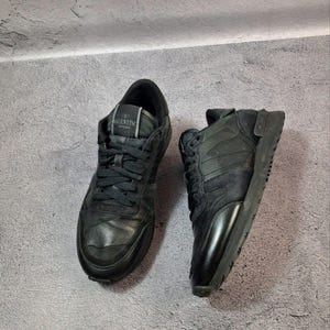 May include: Pair of black sneakers with Valentino branding on the tongue. The shoes feature a mix of textures, including leather and suede, and have black laces. The soles are also black, creating a monochromatic look.