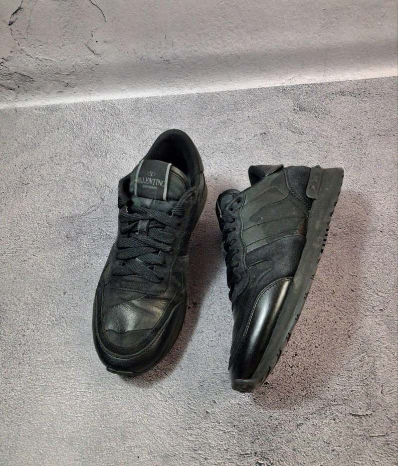 May include: Pair of black sneakers with Valentino branding on the tongue. The shoes feature a mix of textures, including leather and suede, and have black laces. The soles are also black, creating a monochromatic look.