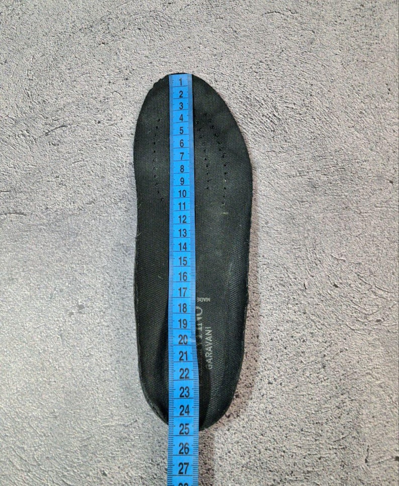 May include: A black shoe insole with a blue measuring tape alongside it. The insole is approximately 11 inches long. The insole has small perforations and the text "GARAVANI" is visible. The background is a textured gray surface.