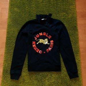 May include: Black sweatshirt with a crew neck and long sleeves. The front features a circular design with the text "JUNGLE - PARIS" in orange, surrounding a tiger graphic. The brand name "KENZO" is also visible.