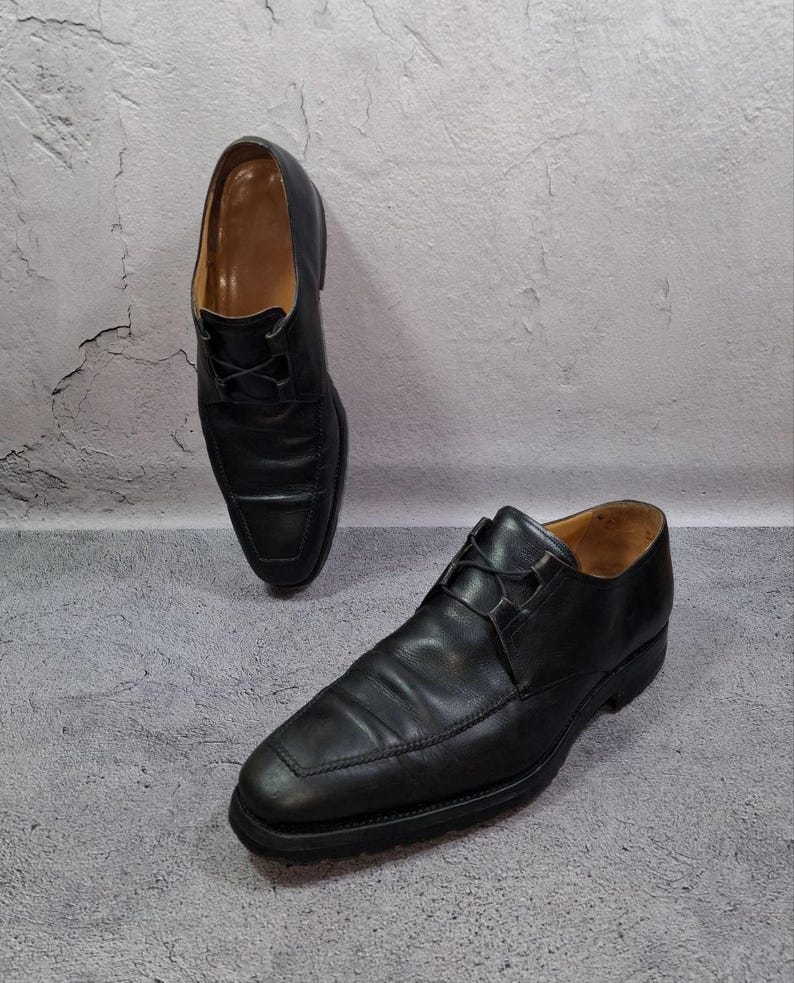 Op de afbeelding: Twee zwarte leren herenschoenen met veters. De schoenen hebben een vierkante neus en staan tegen een grijze achtergrond. De schoenen zijn een klassieke stijl, geschikt voor formele gelegenheden.