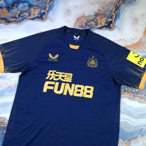 K&ouml;nnte beinhalten: Ein marineblaues Fu&szlig;balltrikot mit gelben Akzenten an &Auml;rmeln und Kragen. Das Trikot zeigt Logos von Castore, FUN88 und ein Vereinswappen. Es hat einen V-Ausschnitt und besteht aus atmungsaktivem Stoff.