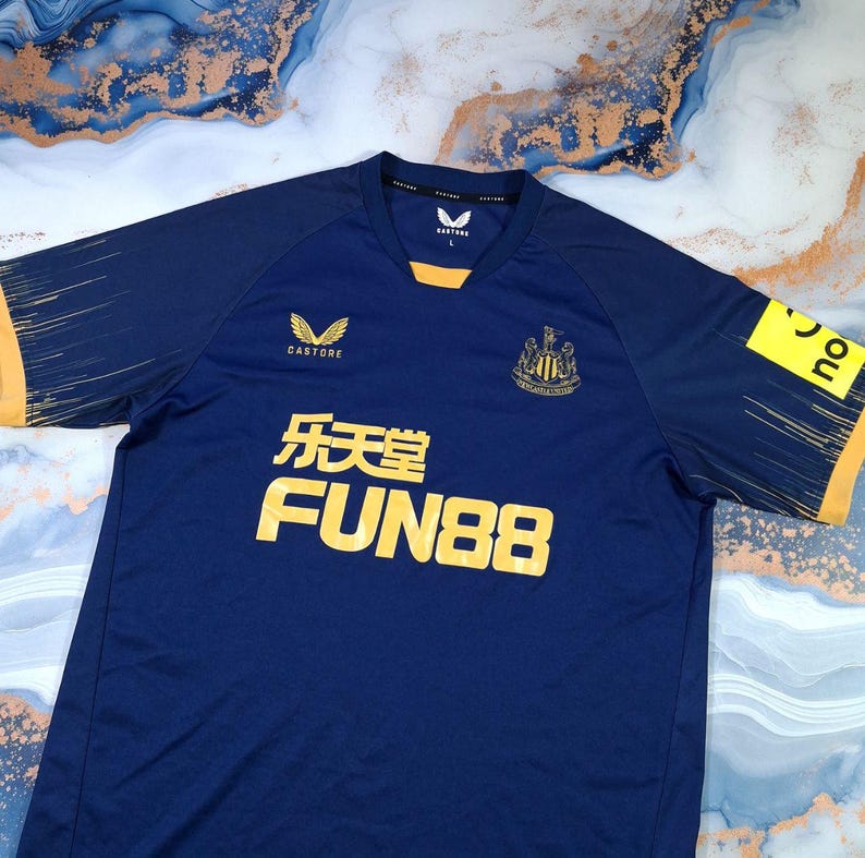 K&ouml;nnte beinhalten: Ein marineblaues Fu&szlig;balltrikot mit gelben Akzenten an &Auml;rmeln und Kragen. Das Trikot zeigt Logos von Castore, FUN88 und ein Vereinswappen. Es hat einen V-Ausschnitt und besteht aus atmungsaktivem Stoff.
