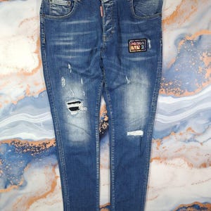 May include: Blue denim jeans with distressed details, including rips and fading. Features a small patch with the text "DSQUARED2" and a red tag. The jeans have a classic five-pocket design.