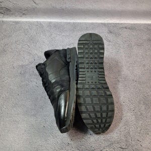 May include: Black sneakers with a textured sole and a combination of leather and suede materials. The sneakers are shown from the side and bottom, showcasing the design and construction. The shoes are on a gray surface.