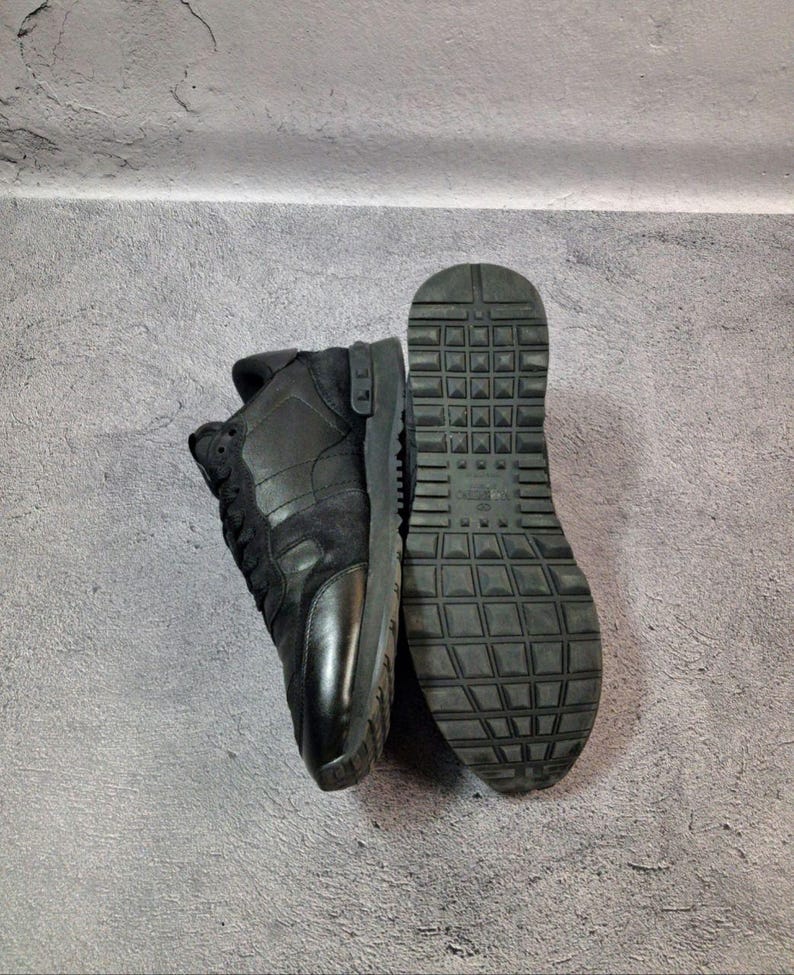 May include: Black sneakers with a textured sole and a combination of leather and suede materials. The sneakers are shown from the side and bottom, showcasing the design and construction. The shoes are on a gray surface.
