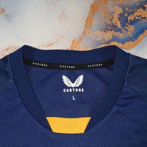 K&ouml;nnte beinhalten: Nahaufnahme eines marineblauen Sporttrikots mit gelbem Detail am Ausschnitt. Der Kragen tr&auml;gt den Markennamen "Castore" und ein wei&szlig;es Logo. Das Trikot ist mit der Gr&ouml;&szlig;e "L" gekennzeichnet. Der Stoff ist glatt, wahrscheinlich synthetisch.