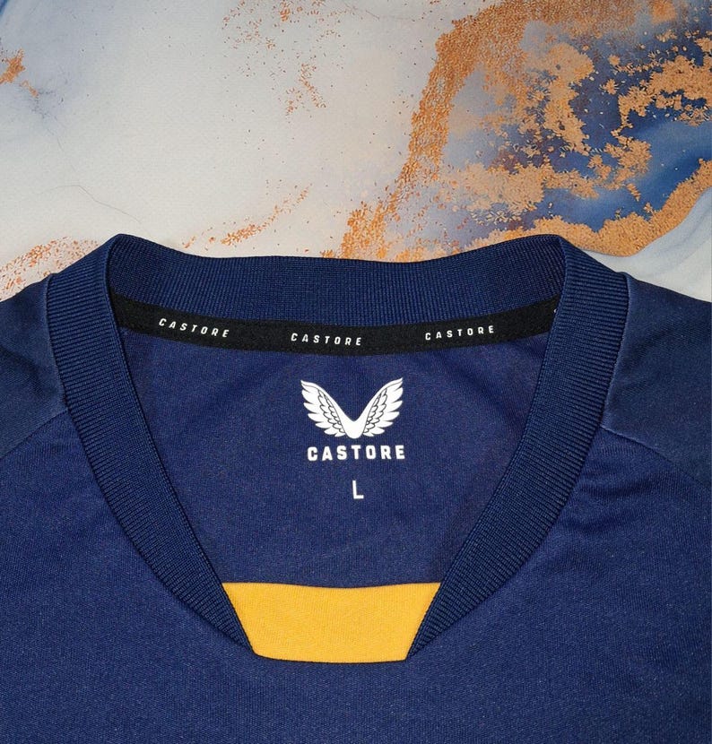 K&ouml;nnte beinhalten: Nahaufnahme eines marineblauen Sporttrikots mit gelbem Detail am Ausschnitt. Der Kragen tr&auml;gt den Markennamen "Castore" und ein wei&szlig;es Logo. Das Trikot ist mit der Gr&ouml;&szlig;e "L" gekennzeichnet. Der Stoff ist glatt, wahrscheinlich synthetisch.