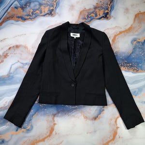 Può includere: Un blazer nero con chiusura a un bottone e revers a lancia. La giacca ha maniche lunghe e una vestibilità sartoriale. La fodera interna è nera. Il blazer è esposto su una superficie marmorizzata.