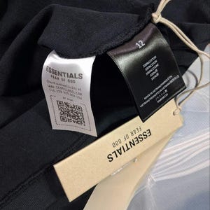 May include: Close-up of a black garment with multiple tags. The tags include the brand name "ESSENTIALS FEAR OF GOD", a QR code, and text indicating the item is 100% cotton and made in Vietnam. The number "12" is also visible.