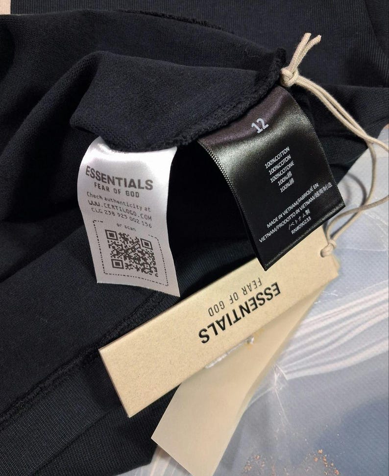 May include: Close-up of a black garment with multiple tags. The tags include the brand name "ESSENTIALS FEAR OF GOD", a QR code, and text indicating the item is 100% cotton and made in Vietnam. The number "12" is also visible.