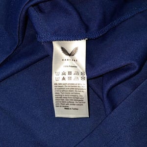 K&ouml;nnte beinhalten: Nahaufnahme eines marineblauen Stoffes mit einem wei&szlig;en Etikett. Das Etikett zeigt den Markennamen "Castore" und Pflegehinweise. Der Stoff besteht aus 100 % Polyester. Auf dem Etikett steht auch "Made in Turkey".