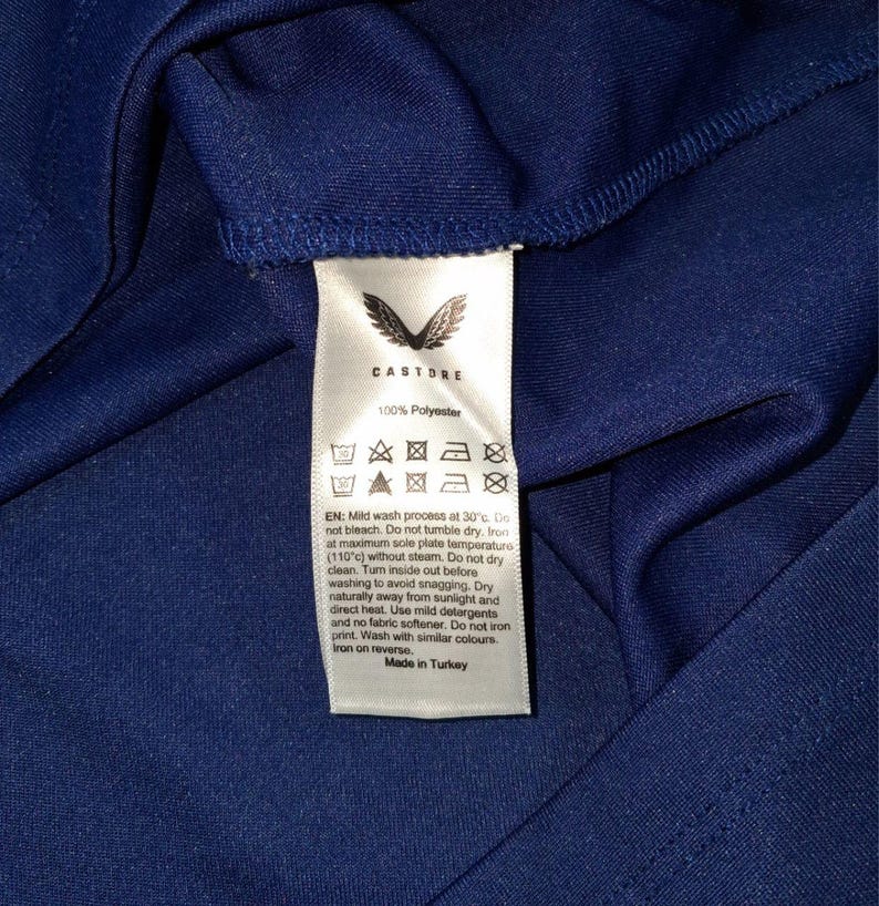 K&ouml;nnte beinhalten: Nahaufnahme eines marineblauen Stoffes mit einem wei&szlig;en Etikett. Das Etikett zeigt den Markennamen "Castore" und Pflegehinweise. Der Stoff besteht aus 100 % Polyester. Auf dem Etikett steht auch "Made in Turkey".