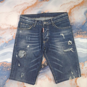 May include: Blue denim shorts with distressed details, including rips and patches. The shorts have a button fly and classic five-pocket styling. A small red tag is visible near the button closure. The shorts are laid flat.