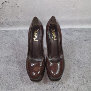 May include: A pair of brown patent leather high-heeled pumps. The shoes have a platform sole and a high heel. The brand logo is visible in gold on the inner side of each shoe.