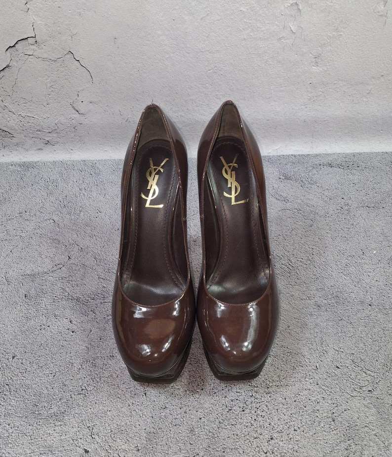 May include: A pair of brown patent leather high-heeled pumps. The shoes have a platform sole and a high heel. The brand logo is visible in gold on the inner side of each shoe.