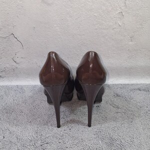 May include: A pair of brown patent leather high-heeled shoes. The shoes have a pointed toe and a stiletto heel. The heels are approximately 4 inches tall. The shoes are set against a neutral background.