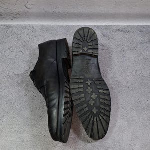 Op de afbeelding: Zwarte leren enkellaarsjes met een profielzool, &eacute;&eacute;n staat rechtop en de andere toont de zool. De laarzen hebben een klassiek ontwerp met een ronde neus en een stevige, getextureerde zool voor grip.