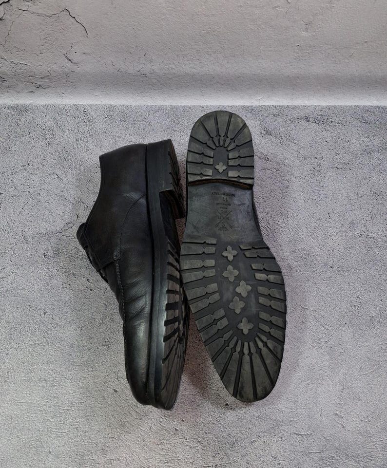 Op de afbeelding: Zwarte leren enkellaarsjes met een profielzool, &eacute;&eacute;n staat rechtop en de andere toont de zool. De laarzen hebben een klassiek ontwerp met een ronde neus en een stevige, getextureerde zool voor grip.