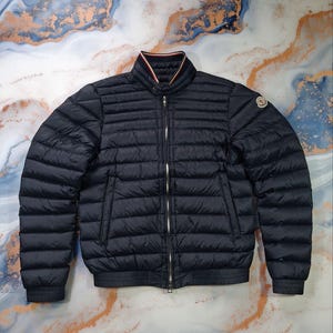 Puede incluir: Chaqueta acolchada azul marino con cremallera completa y cuello acanalado. La chaqueta tiene un pequeño logotipo en la manga superior izquierda y un dobladillo y puños acanalados. El cuello tiene rayas rojas y blancas.