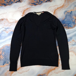 May include: A black V-neck jumper with long sleeves. The jumper features a small logo near the chest and a brand label at the neckline. The fabric appears to be a fine knit. The jumper is displayed on a patterned surface.