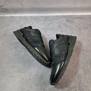 May include: A pair of black sneakers with a combination of suede and leather materials. The shoes have a low-profile design with a lace-up closure and a thick, textured sole. The sneakers are displayed against a gray background.