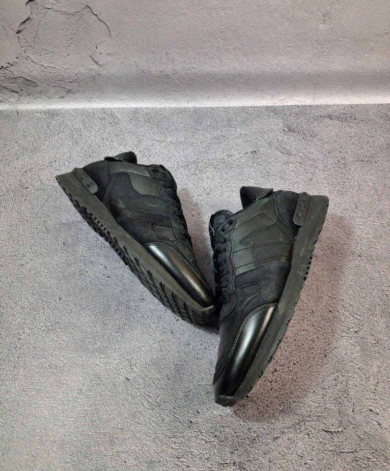 May include: A pair of black sneakers with a combination of suede and leather materials. The shoes have a low-profile design with a lace-up closure and a thick, textured sole. The sneakers are displayed against a gray background.