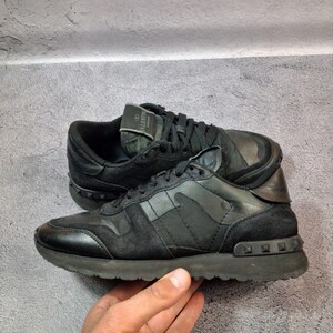 May include: Black sneakers with a camouflage design on the side panels. The shoes have a suede and leather construction, with a studded detail on the heel and sole. The laces and soles are also black.