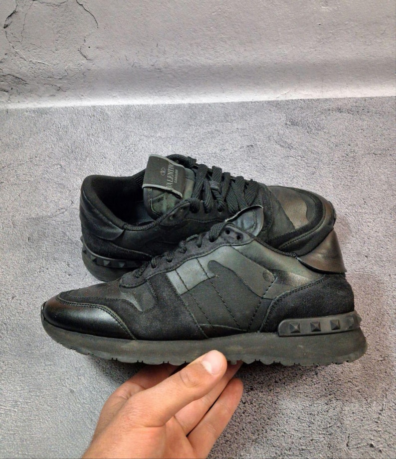 May include: Black sneakers with a camouflage design on the side panels. The shoes have a suede and leather construction, with a studded detail on the heel and sole. The laces and soles are also black.