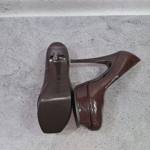 May include: A pair of brown platform high heels. The shoes have a glossy finish, a square toe, and a thin stiletto heel. The platform sole adds height and a modern touch to the classic design.