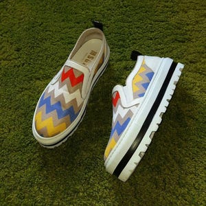 May include: Pair of white slip-on sneakers with a colorful zigzag pattern in red, yellow, blue, and brown. The shoes have a thick white sole with a black stripe and a black pull tab.