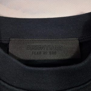 May include: Close-up of a navy blue crew neck sweatshirt. The black label on the collar features the embossed text "ESSENTIALS" above "FEAR OF GOD". The fabric has a ribbed texture.