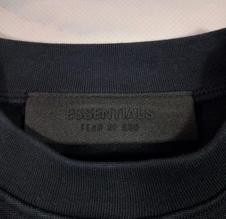 May include: Close-up of a navy blue crew neck sweatshirt. The black label on the collar features the embossed text "ESSENTIALS" above "FEAR OF GOD". The fabric has a ribbed texture.