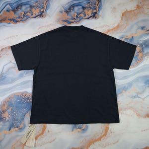 May include: A black t-shirt with a tag attached. The shirt is laid flat, showcasing its simple design. The tag reads "ESSENTIALS". The fabric appears to be a soft, comfortable material. The background has a marbled pattern.