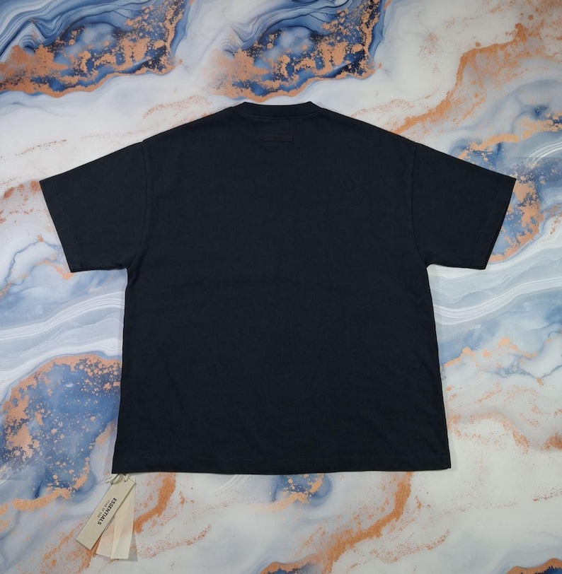 May include: A black t-shirt with a tag attached. The shirt is laid flat, showcasing its simple design. The tag reads "ESSENTIALS". The fabric appears to be a soft, comfortable material. The background has a marbled pattern.