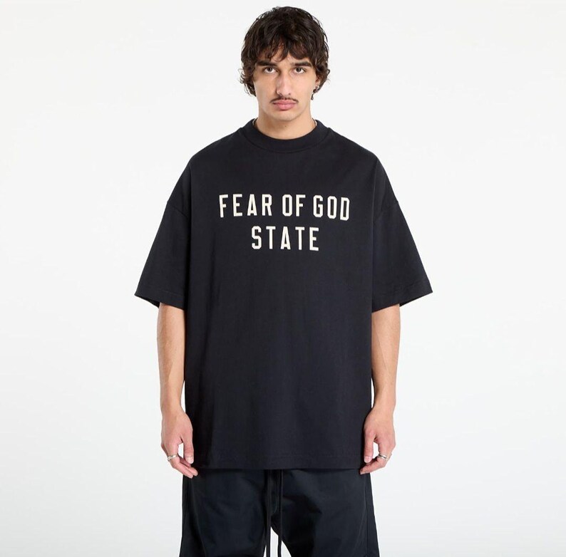 May include: Black t-shirt with the text "FEAR OF GOD STATE" in white block letters. The shirt has short sleeves and a crew neck. The model is wearing black pants.