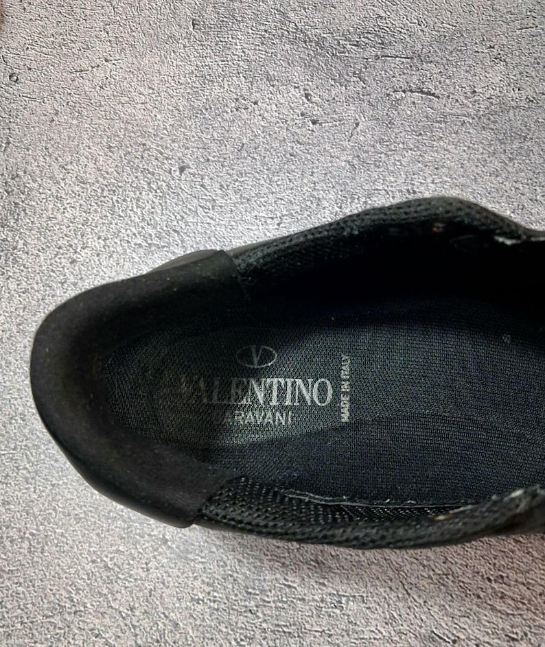 May include: Close-up of a black shoe interior. The shoe features the brand name "VALENTINO GARAVANI" in white lettering. The words "MADE IN ITALY" are also visible. The shoe appears to be made of a mesh material.
