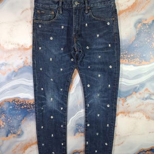 Op de afbeelding: Donkerblauwe denim jeans met een normale pasvorm. De jeans zijn versierd met een herhalend patroon van witte schedel- en kroonemblemen. De jeans hebben een klassiek ontwerp met vijf zakken en een knoop- en ritssluiting.