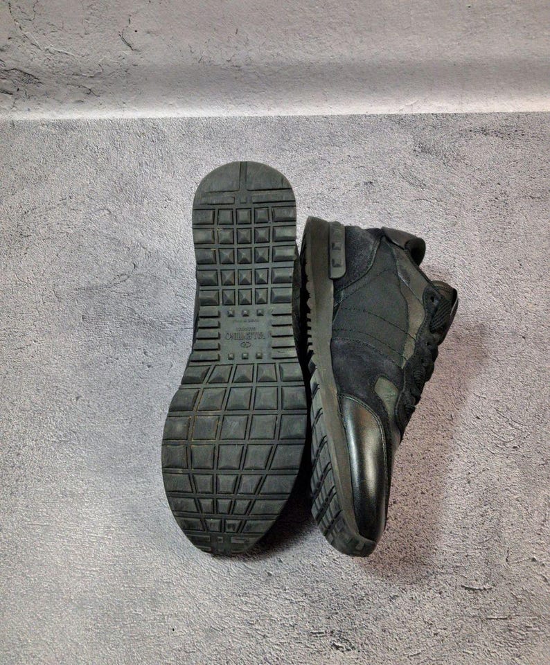 May include: Black sneakers with a textured sole and a combination of suede and leather panels. The sole is thick and features a grid-like pattern. The sneakers are displayed against a gray background.