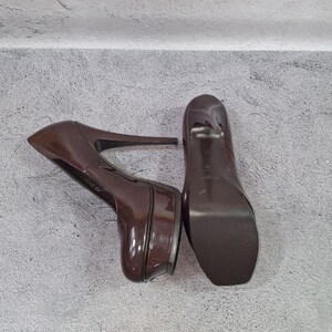 May include: Pair of glossy, brown platform high heels. The shoes have a rounded toe and a stiletto heel. The platform sole adds height. The heels are positioned on a gray surface.