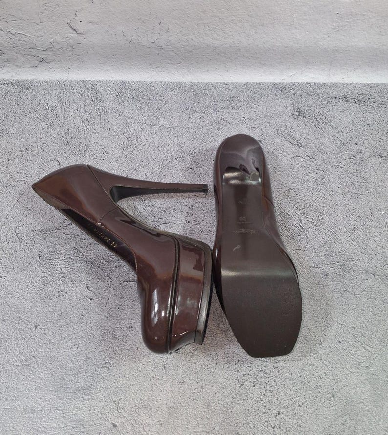 May include: Pair of glossy, brown platform high heels. The shoes have a rounded toe and a stiletto heel. The platform sole adds height. The heels are positioned on a gray surface.