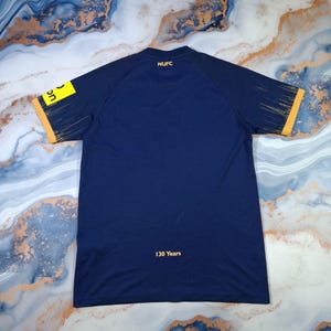 K&ouml;nnte beinhalten: Marineblaues Sporttrikot mit gelben Akzenten an &Auml;rmeln und Kragen. Das Trikot hat den Aufdruck "NUFC" auf dem R&uuml;cken, darunter der Text "130 Years". Auf dem linken &Auml;rmel befindet sich ein gelbes Rechteck mit Text.