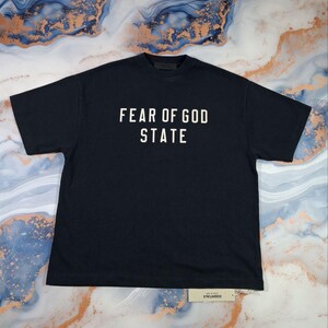 May include: Black t-shirt with the text "FEAR OF GOD STATE" in white block letters. The shirt is laid flat, showing the front and the short sleeves. A small tag is visible at the bottom.