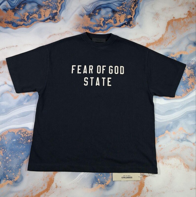 May include: Black t-shirt with the text "FEAR OF GOD STATE" in white block letters. The shirt is laid flat, showing the front and the short sleeves. A small tag is visible at the bottom.