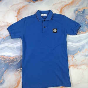 May include: A short-sleeved, cobalt blue polo shirt with a black collar and sleeve trim. The shirt features a two-button placket and a small square patch on the chest. The shirt is made of a soft, breathable fabric.