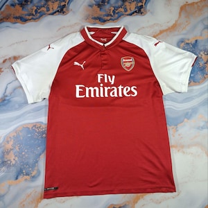 May include: A red and white Arsenal soccer jersey. The jersey features the Arsenal team crest, the Puma logo, and the "Fly Emirates" sponsor logo. The sleeves are white, and the body is red. The jersey has a collared neckline.