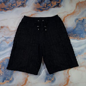 Puede incluir: Shorts negros con un patrón geométrico. Los shorts tienen una cintura con cordón y punteras metálicas plateadas. La tela tiene una apariencia texturizada, similar a la felpa. Los shorts son hasta la rodilla.