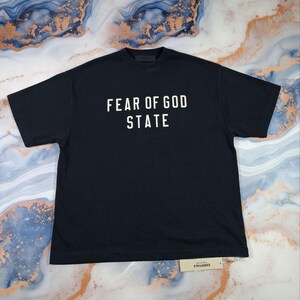 May include: A navy blue t-shirt with the words "FEAR OF GOD STATE" printed in white block letters. The shirt has a crew neck and short sleeves. A small tag is attached to the bottom of the shirt.